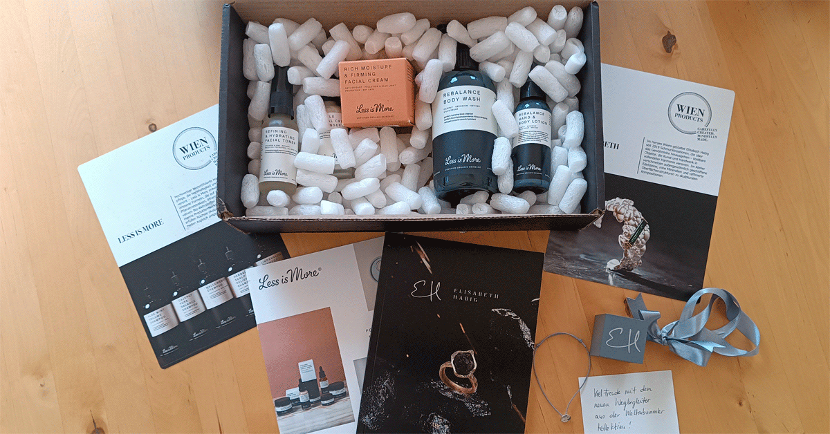 Surprise package with travel-size skincare by Less is More and a pendant from the Weltenbummler Collection by Elisabeth Habig, displayed with brochures, ribbon, and a handwritten note on a wooden table.