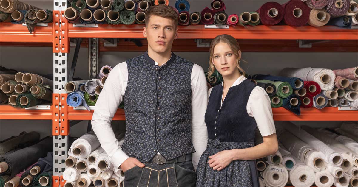A man in a jacquard vest and lederhosen and a woman in a matching dirndl model traditional clothing from the Felicitas & Hans Fall/Winter 2025 collection, posing in front of shelves filled with fabric rolls.