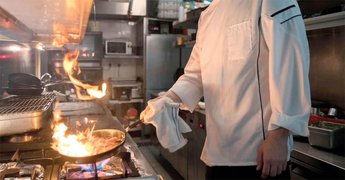 Chef in white jacket flambéing food in a pan over a gas stove in a professional kitchen
