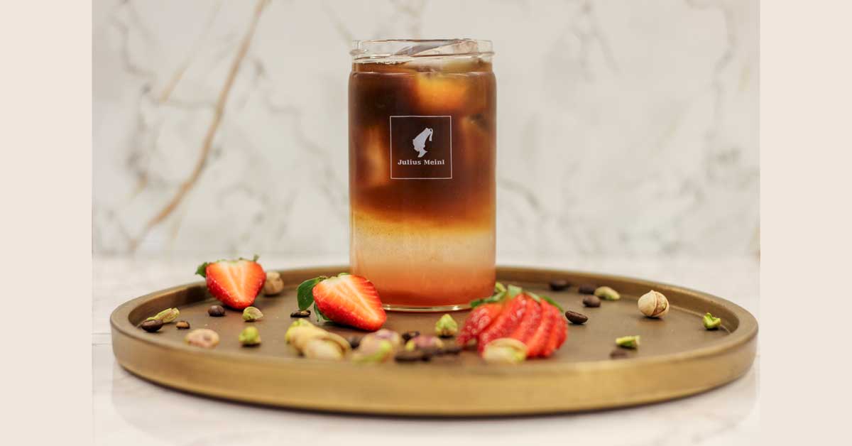 Glass of Strawberry-Pistachio Espresso Tonic with Julius Meinl logo, surrounded by strawberries and pistachios.