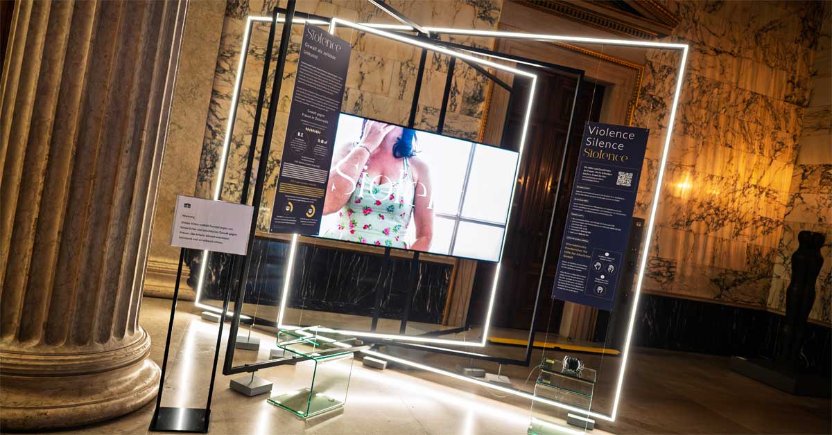 'Siolence' exhibition setup in Parliament: lit metal frames, info boards and a video of an anonymised woman, placed in a marble hall.
