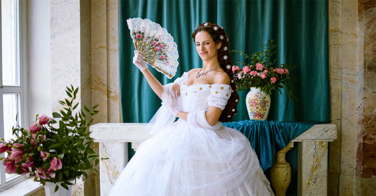 Campaign image 'Just Sisi' from the 'Lebensgefühl Österreich' campaign, representing Empress Elisabeth of Austria (Sisi); Photo: ÖW/Linda Schäffler. A woman in a white historical gown with a fan, set against a teal curtain in a grand interior.
