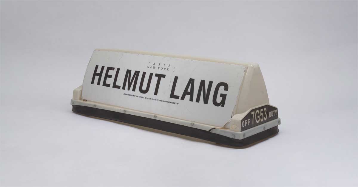 White New York City taxi roof sign displaying the black HELMUT LANG logo with the words 'Paris New York,' part of the designer’s minimalist advertising campaign from 1998–2004, now in the MAK Helmut Lang Archive.