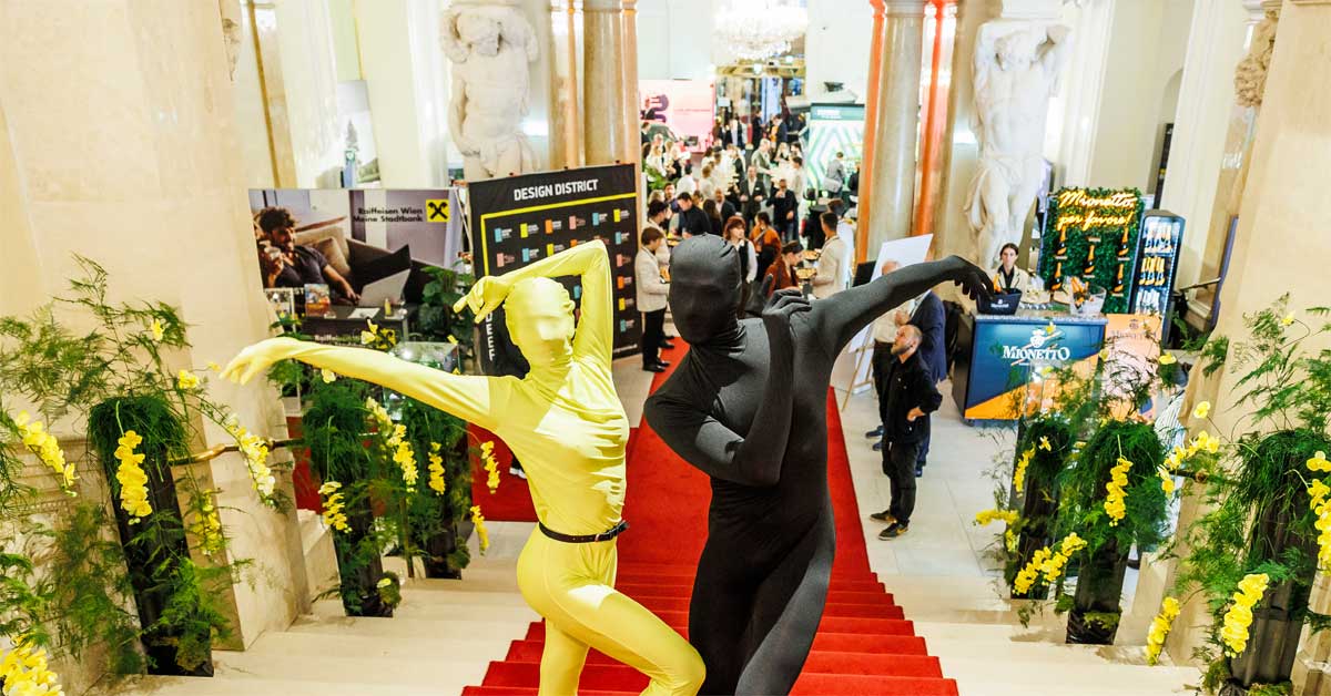 Two performers in full-body costumes pose on the red staircase at the Design District in the Hofburg, surrounded by guests and design installations.