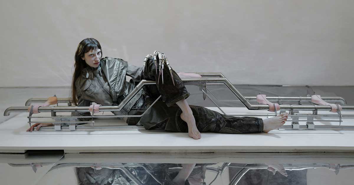 Artist Natália Sýkorová posing barefoot within her metallic installation, wearing avant-garde clothing, her hand transformed into alien-like mechanical prosthesis, reflecting themes of technology and survival.