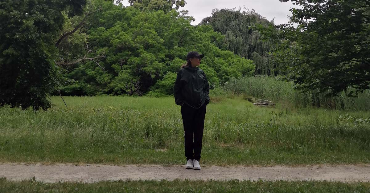 A hiker in a green waterproof jacket, cap, black trousers and sturdy trekking shoes stands on a woodland path.