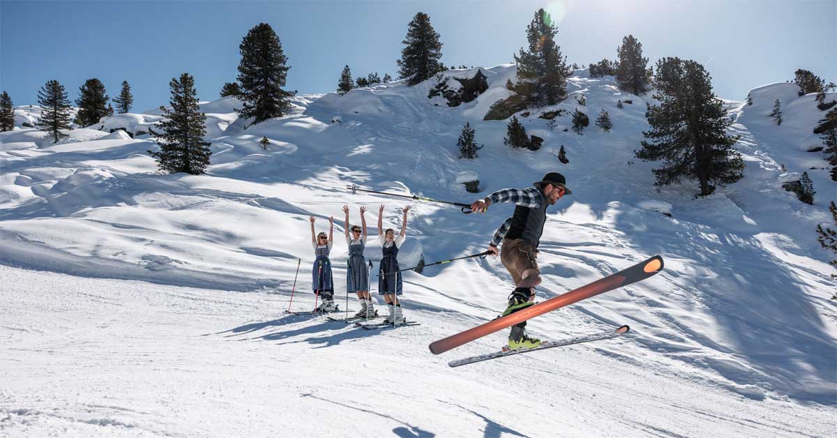 Skier in Lederhose jumping on a sunny slope with women in Dirndl cheering behind him in the Zillertal Arena