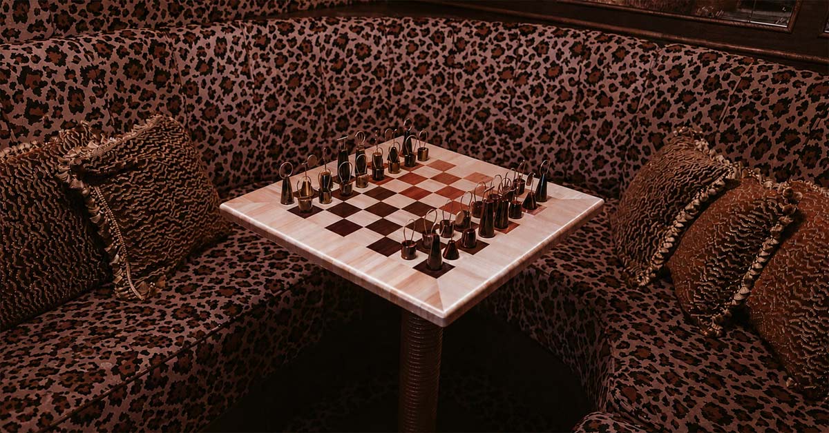 NADÁLI chessboard with sculptural metal chess pieces on a patterned table in a richly decorated lounge interior.