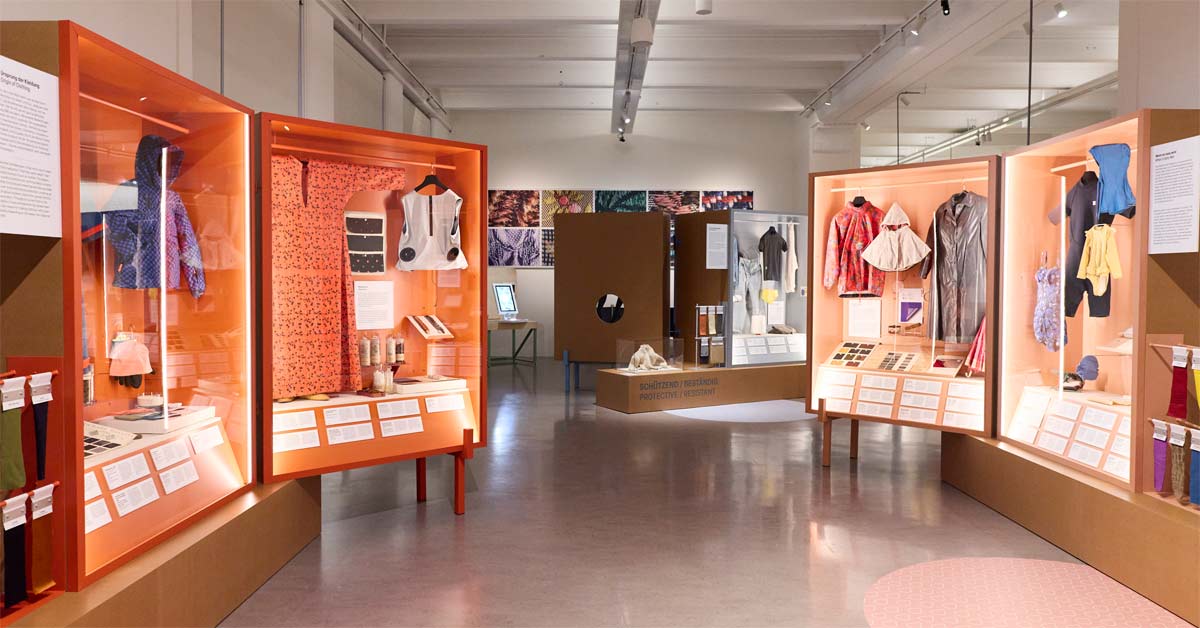Orange-framed cabinets displaying garments and fabric samples at the Tec/xtile exhibition in Vienna.