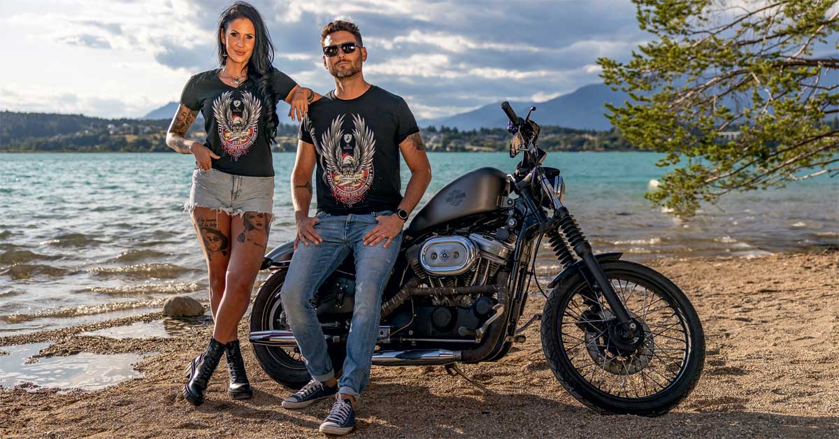 Anita Wagner and Alexander Knapp wearing 2025 Faaker See Biker Shirts beside a Harley-Davidson at Lake Faak, with water, mountains, and sky behind them.