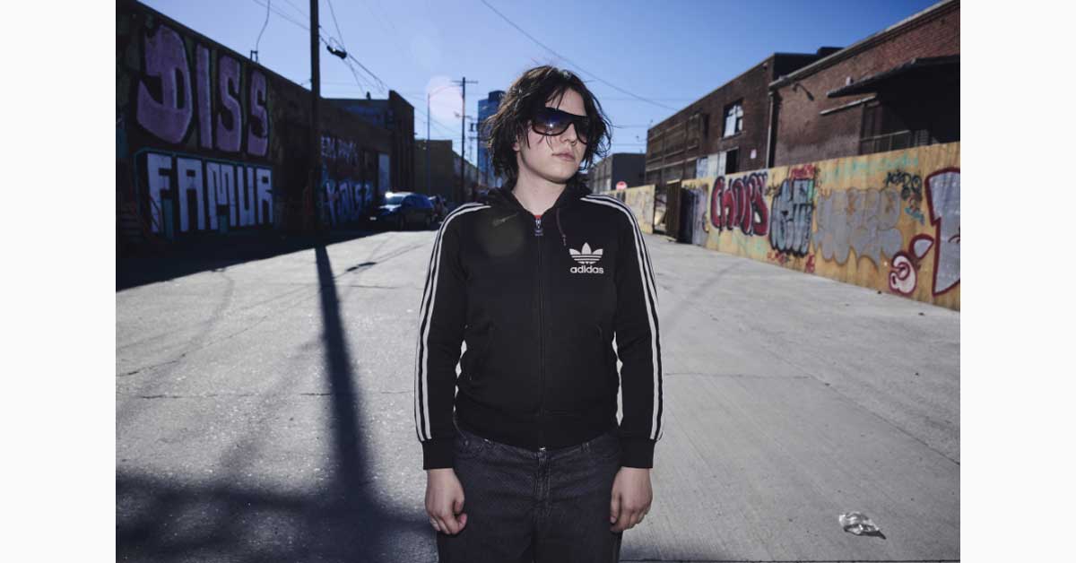 Austrian hyperpop musician Filly standing in an urban street wearing a black sports jacket and sunglasses.